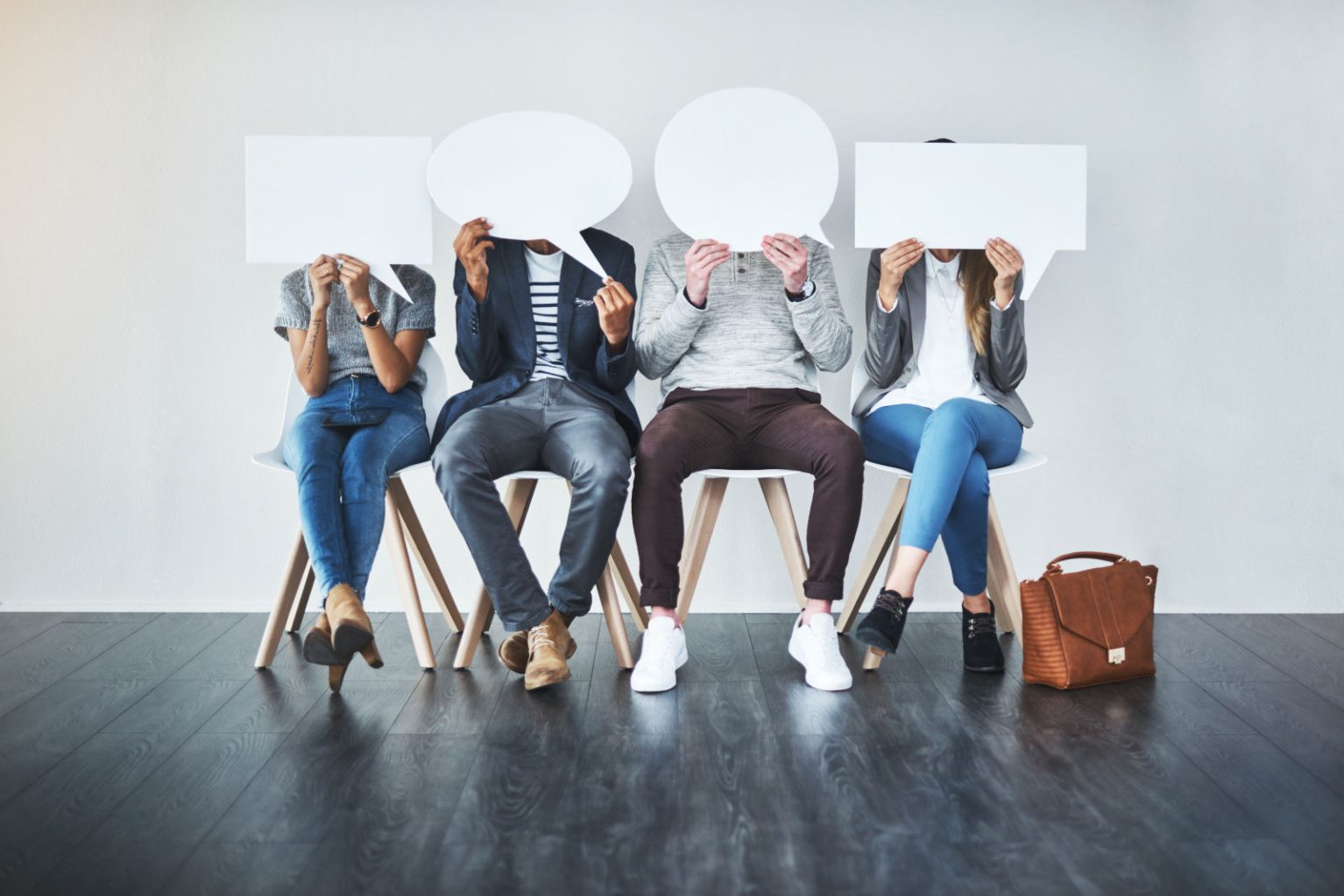 Vier Menschen sitzen auf Stühlen in einer Reihe und halten sich große Sprechblasen vors Gesicht, Symbolbild für Bewerbungen