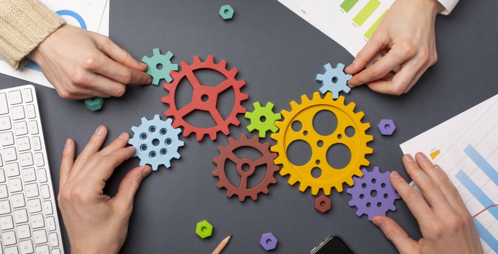 Vier Hände legen auf einem Schreibtisch bunte Zahnräder aneinander - Symbolbild für agiles Projektmanagement im Onlinemarketing und Web Development