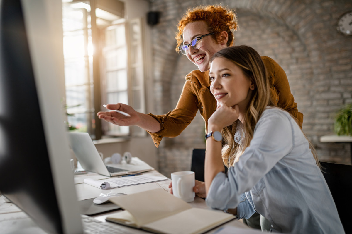 Zwei junge Frauen sitzen vor einen geöffneten Laptop an einem Schreibtisch, Symbolbild für Kundengeschichten im Marketing