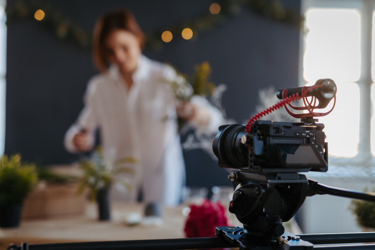 Im Hintergrund sieht man verschwommen eine Frau in weißer Bluse, im Vordergrund ist eine Kamera, die sie aufnimmt, Symbolbild für Kundengeschichten im Marketing.