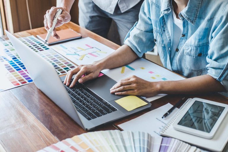 Zwei Menschen sitzen an einem Schreibtisch mit offenem Laptop und diversen Unterlagen